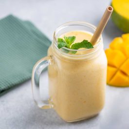 Creamy mango smoothie topped with mint in a glass mug beside sliced mango and a green napkin.