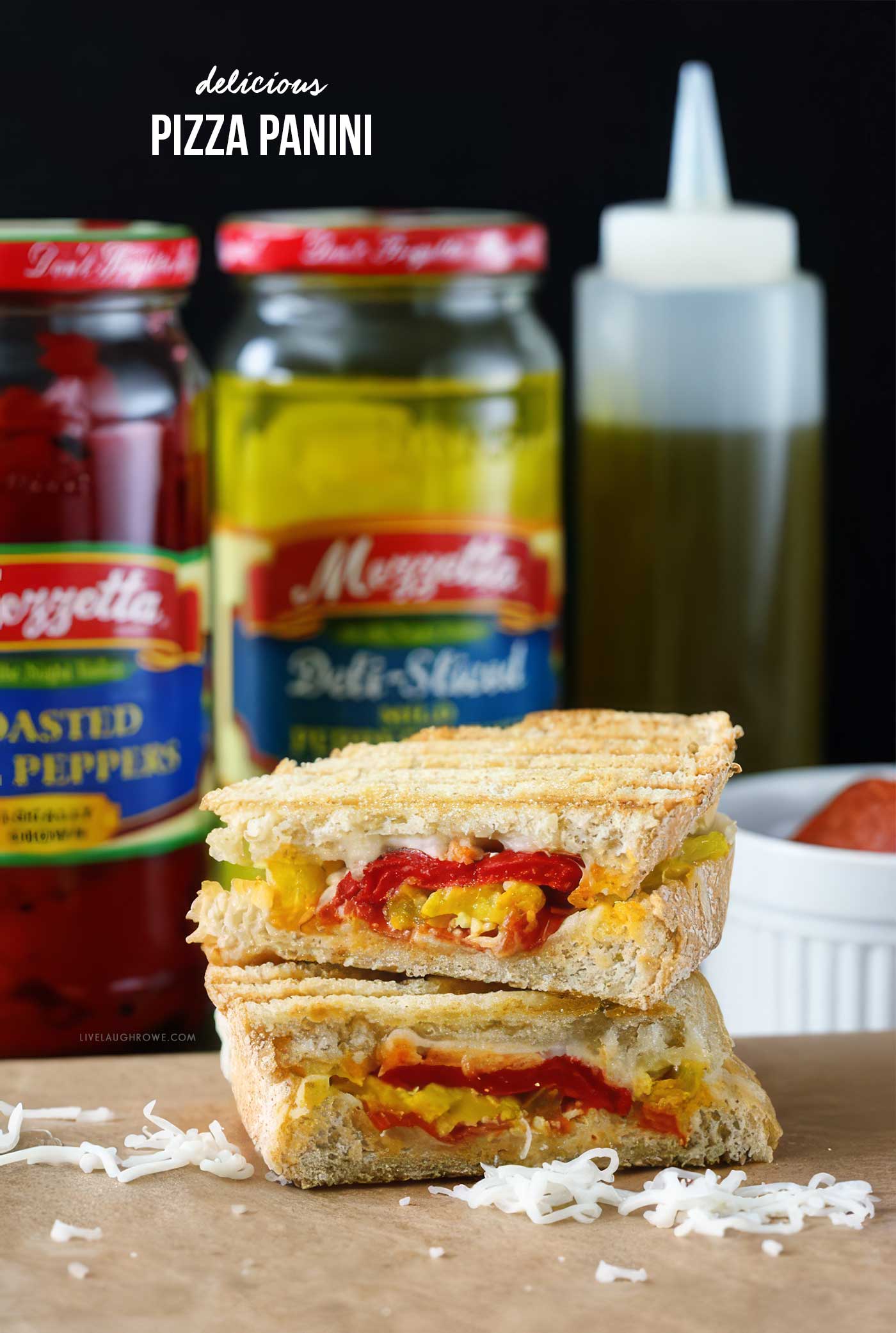 Pizza panini with melted mozzarella, pepperoni, roasted red peppers, and pepper rings stacked in front of jars of pizza toppings.