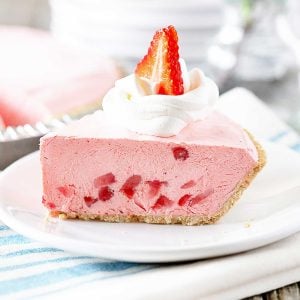 Slice of No Bake Strawberry Jello Pie on a white plate. Pic slice is topped with cool whip and a strawberry slice.
