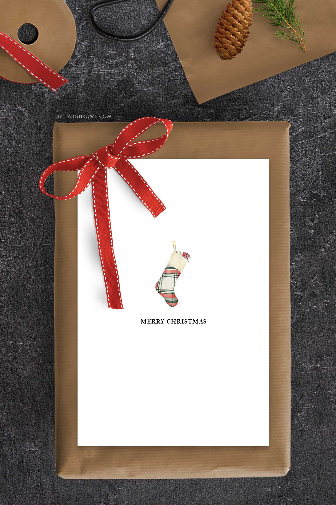 Printable Christmas card with plaid stocking surrounded by kraft paper, scissors, red ribbon, and pinecone.