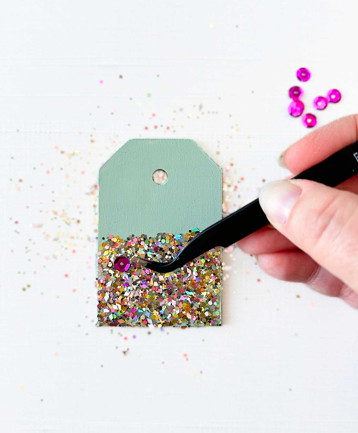 a human hand holding a pair of tweezers and applying a sequin to some glitter