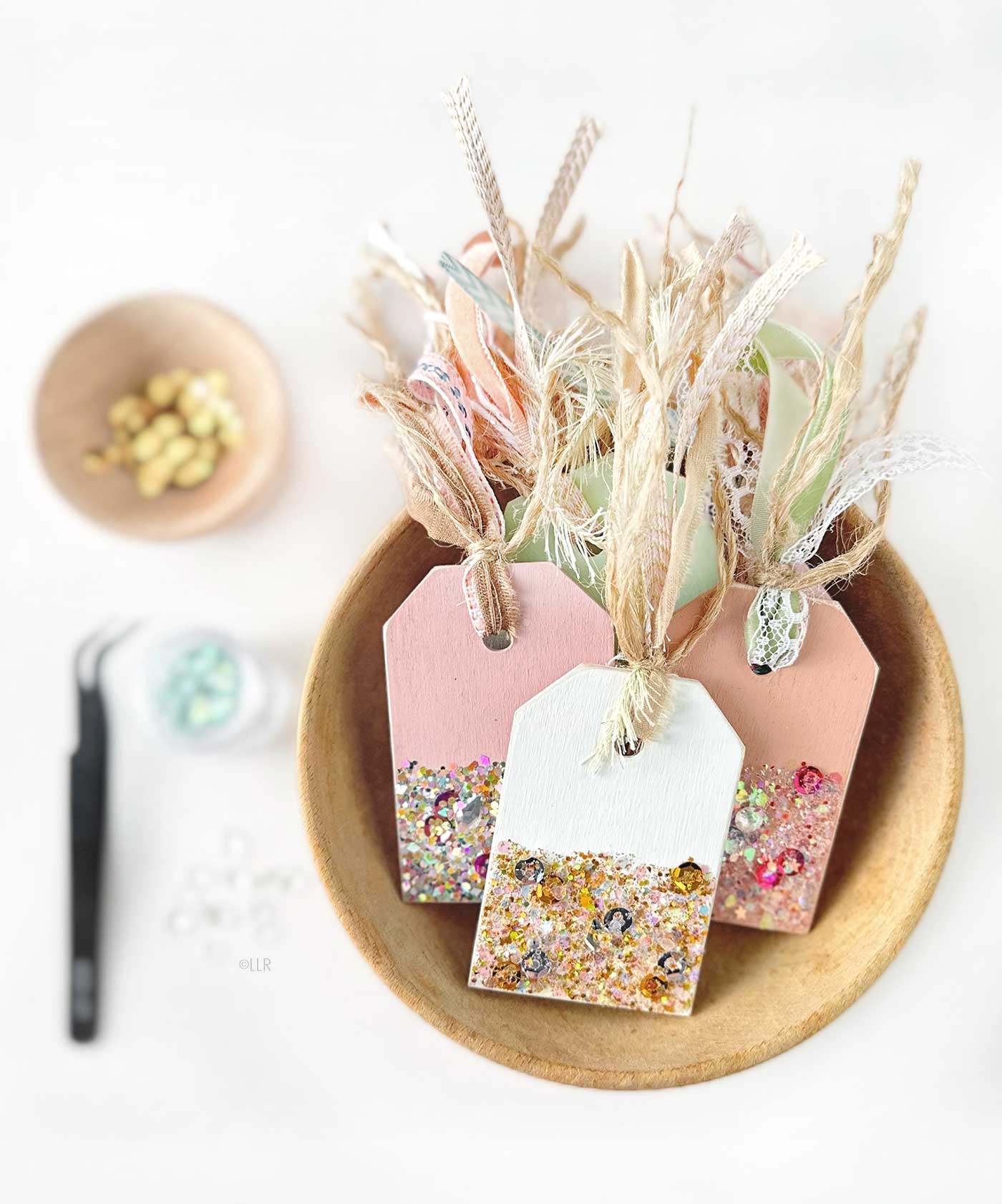 Wood Gift Tags with glitter in a bowl surrounded by other supplies such as sequins, tweezers and glitter