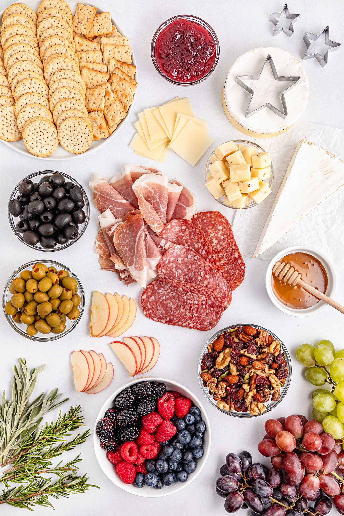 all the ingredients laid out on a white counter for a Christmas charcuterie board. ingredients include crackers, cranberry sauce, salami, fruits, nuts, granola, cheese, honey and more