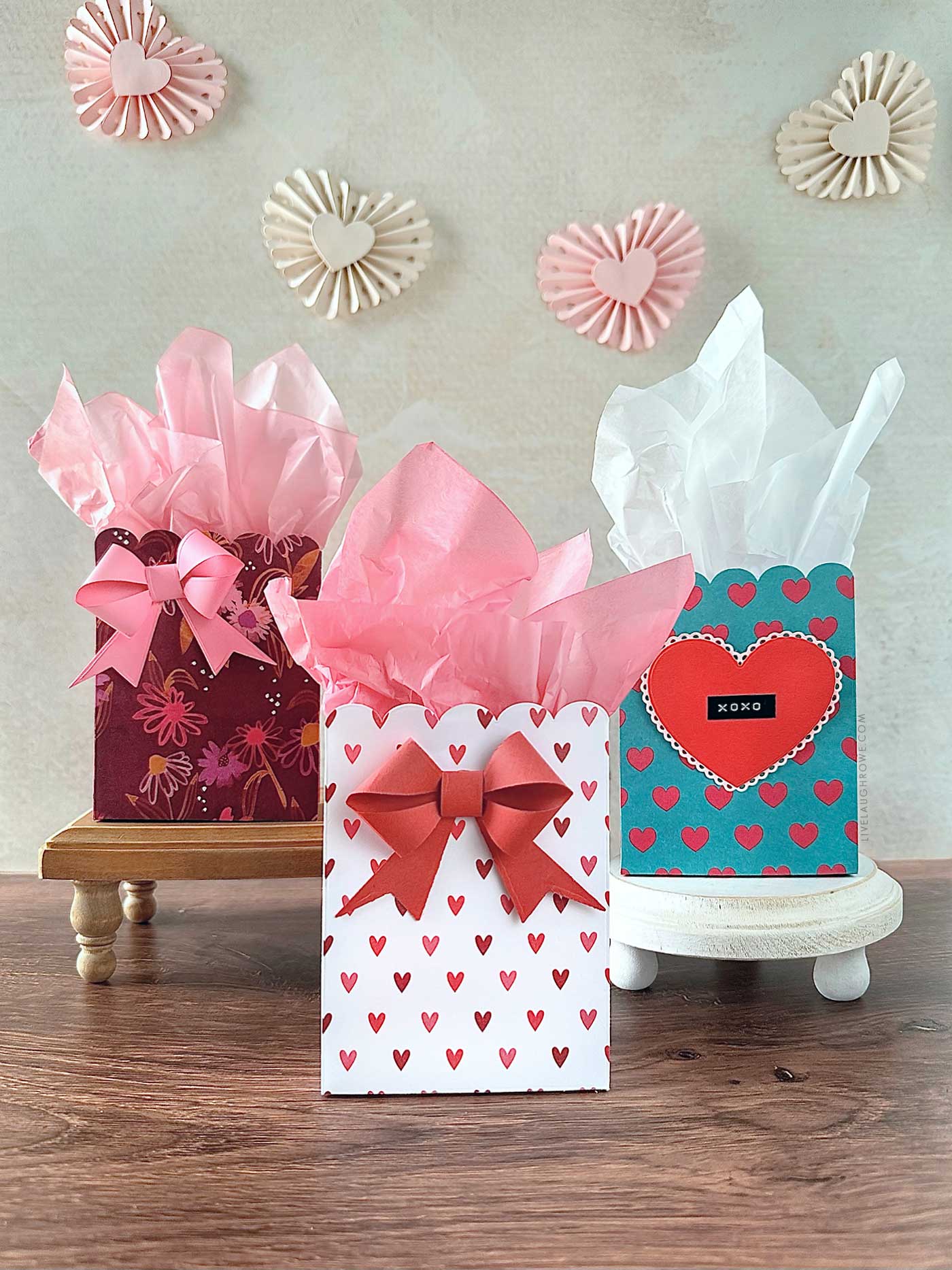 three scalloped paper gift bags with pink and white tissue paper. A heart rosette garland is in the background