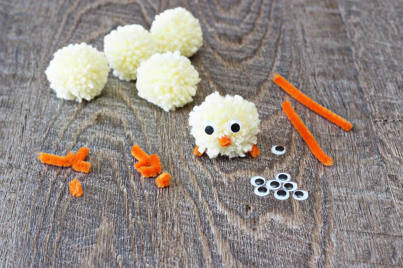 pom pom chick with the supplies to make one around it, such as yellow pom poms, orange pipe cleaner and googly eyes