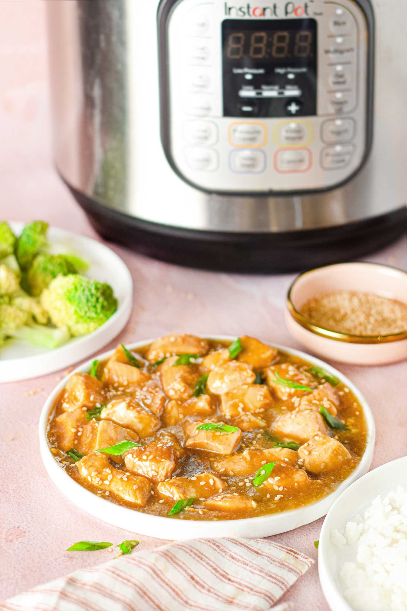 plate with broccoli, a bowl of sesame seeds and a plate full of Teriyaki Chicken with an Instant Pot in the background