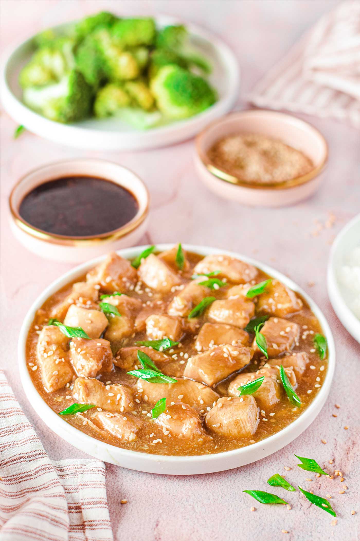 broccolie, bowl of teriyaki sauce and sesame seeds behind a place of chicken in a teriyaki sauce with green onion and sesame seeds sprinkled on it