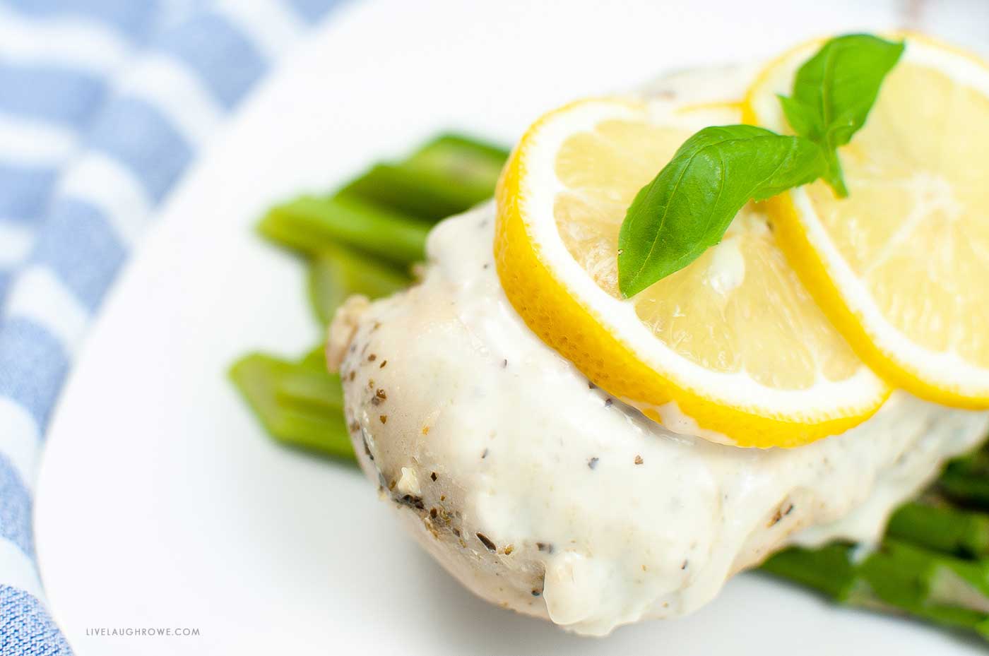 striped napkin with a white place on top of it. The plate contains some asparagus, topped with a chicken breast. On the chicken is a creamy lemon sauce, lemon slices and an herb sprig