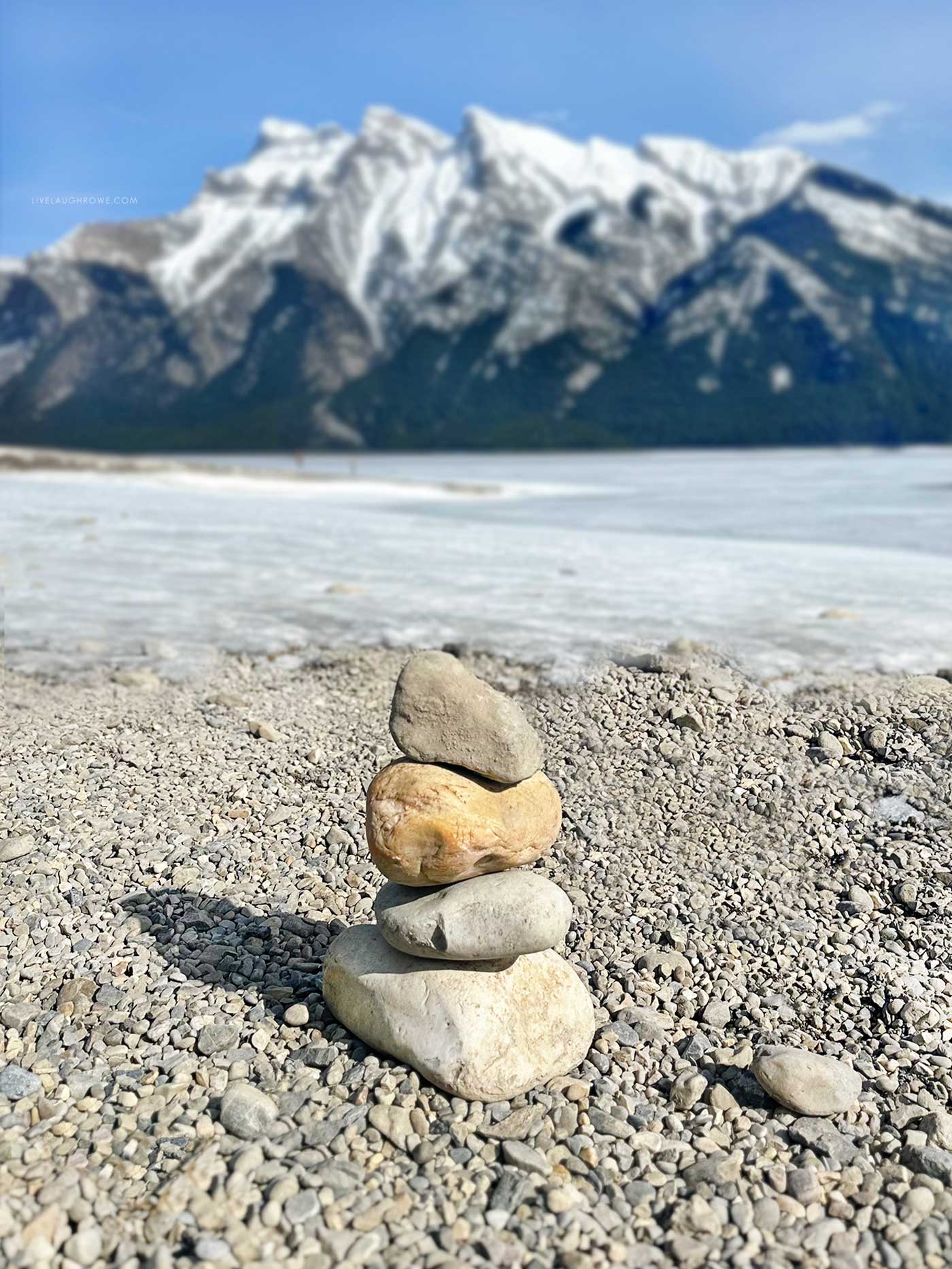 rock statue on a pebble beach in front of lake minnewanka and beautiful mountains in the background
