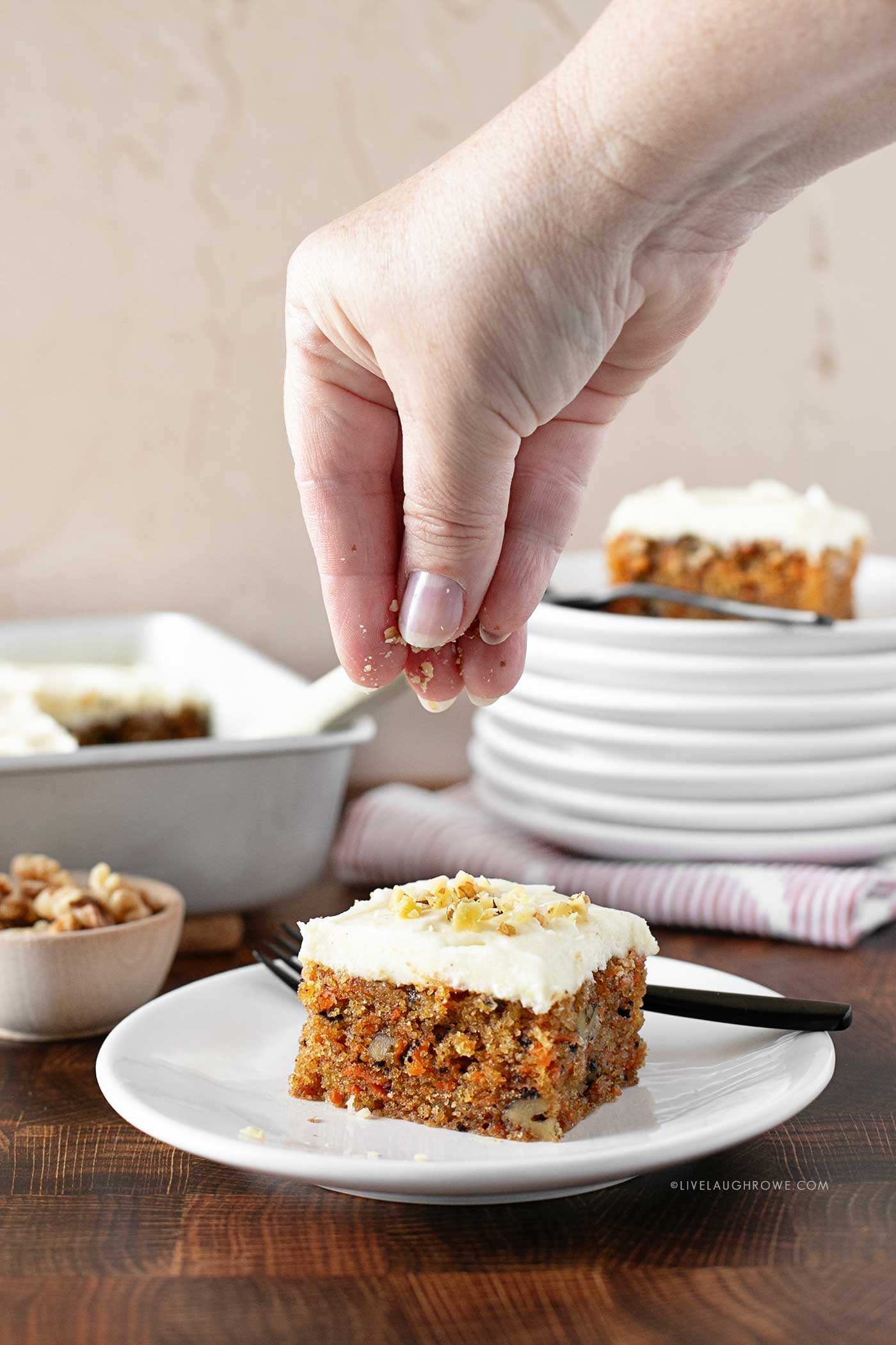 hand sprinkling chopped walnut on a slice of carrot cake sitting on a white place.