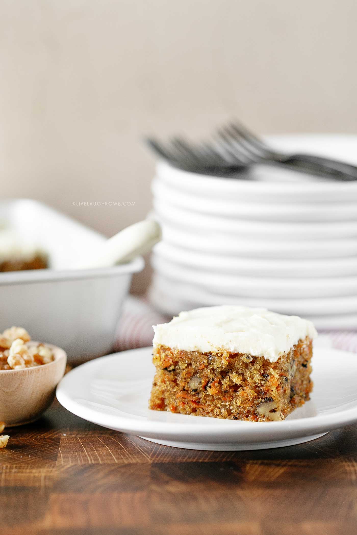 slice of carrot cake with cream cheese frosting on a white plate. Cake and a pile of plates are in the background