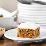 slice of homemade carrot cake with cream cheese frosting on a white plate. Cake and a pile of plates are in the background