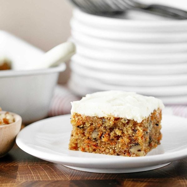 slice of homemade carrot cake with cream cheese frosting on a white plate. Cake and a pile of plates are in the background