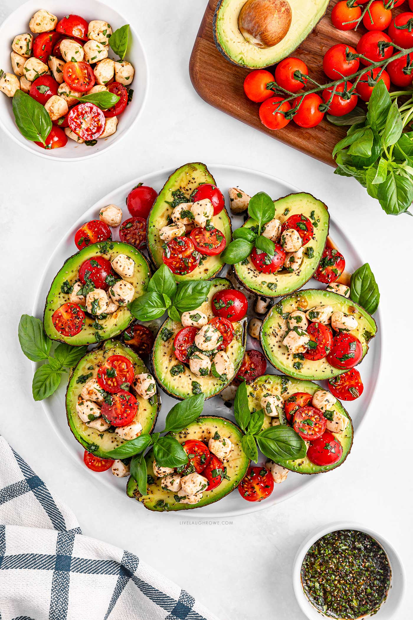 a large whiteround platter full of stuffed avocados on a white background surrounded by ingredients and a hand towel