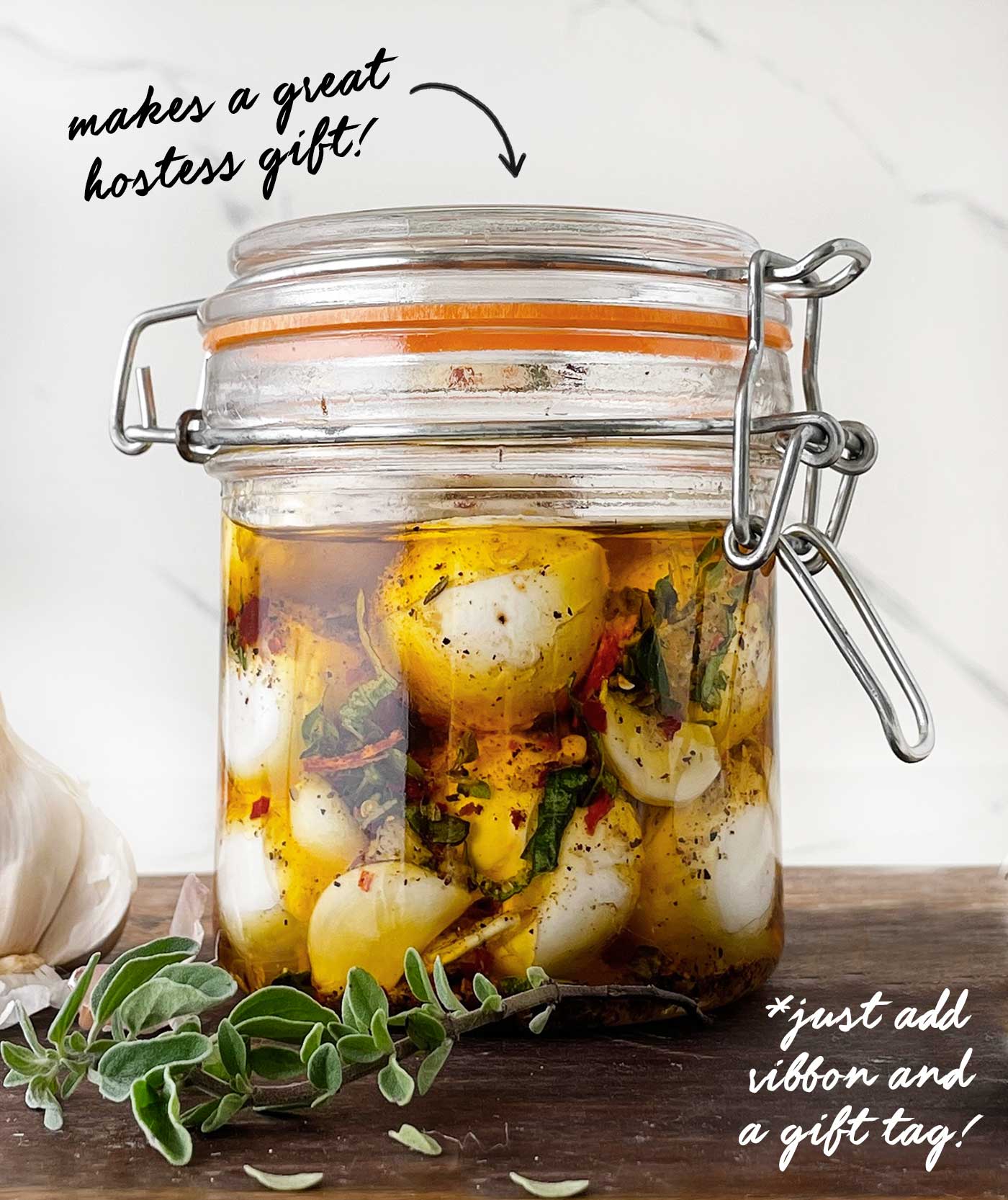 Close-up of fresh mozzarella balls soaking in a flavorful herb and olive oil marinade inside a sealed glass jar. text overlay about this making a great hostess gift.