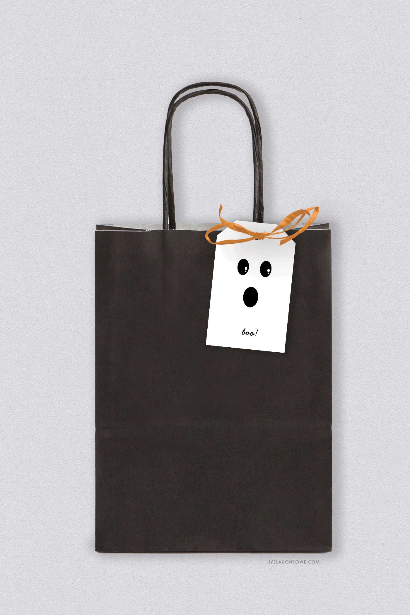 A black paper gift bag decorated with a small white tag featuring a playful ghost face and orange ribbon.