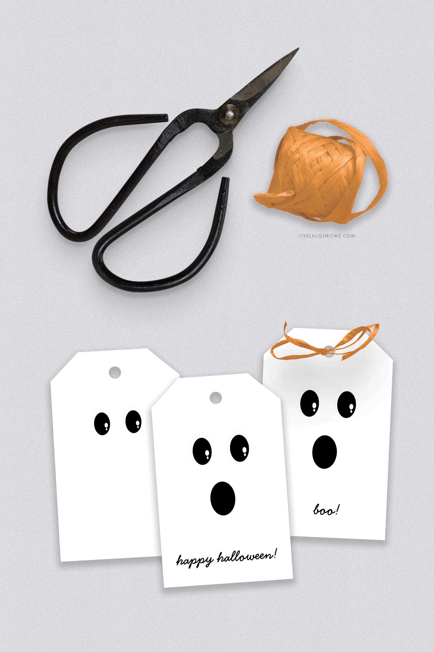 Three finished Halloween tags with cheerful ghost faces, one tied with orange ribbon next to black scissors and a spool of twine.