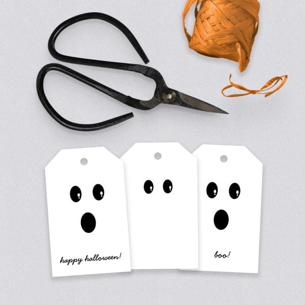 Three finished Halloween tags with cheerful ghost faces next to black scissors and a spool of twine.