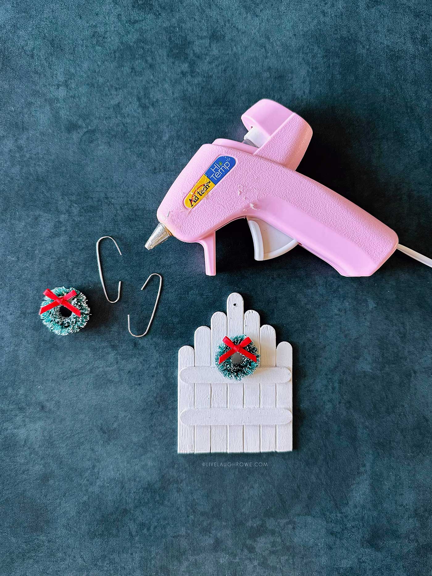 Pink hot glue gun beside mini wreaths and a white scalloped popsicle stick fence ornament ready for embellishing.
