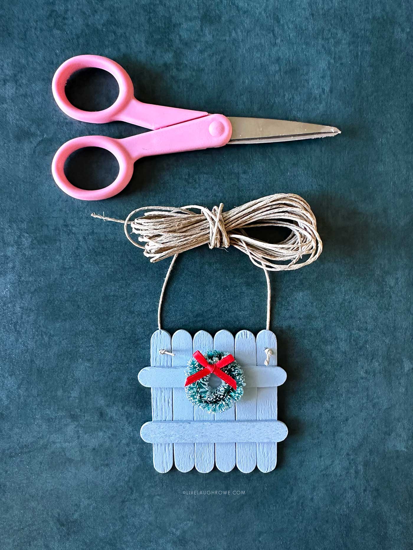 Pink scissors and twine next to a small blue popsicle stick fence ornament decorated with a mini wreath and bow.