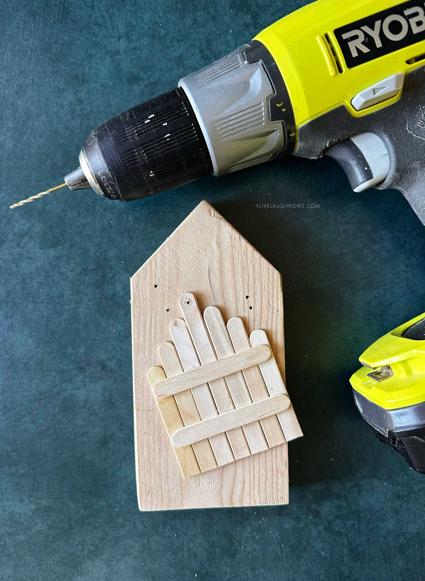 Handheld drill creating holes in a wooden popsicle stick fence ornament for adding a hanger.