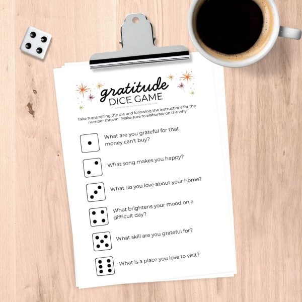 Clipboard displaying the Gratitude Dice Game printable alongside a die and a coffee cup, ready for play.
