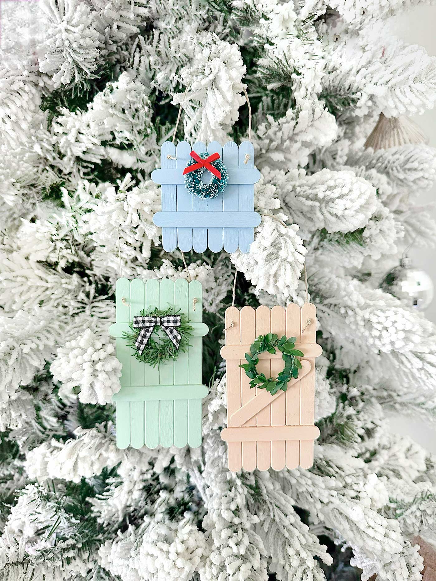 Pastel popsicle stick fence ornaments hanging on a flocked Christmas tree with mini wreath embellishments.
