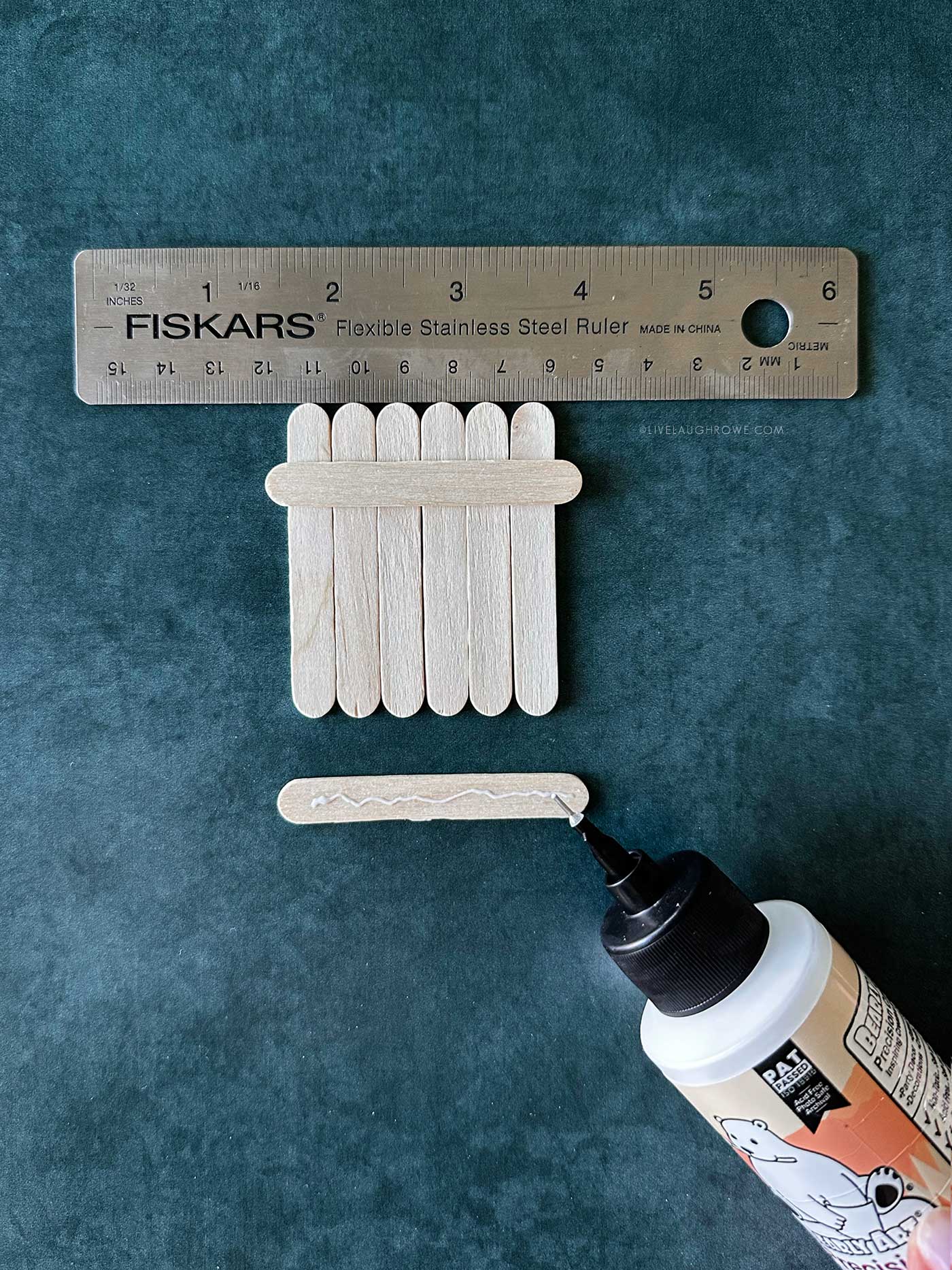 Popsicle sticks aligned with a ruler while glue is applied to a crossbar for assembling ornament.