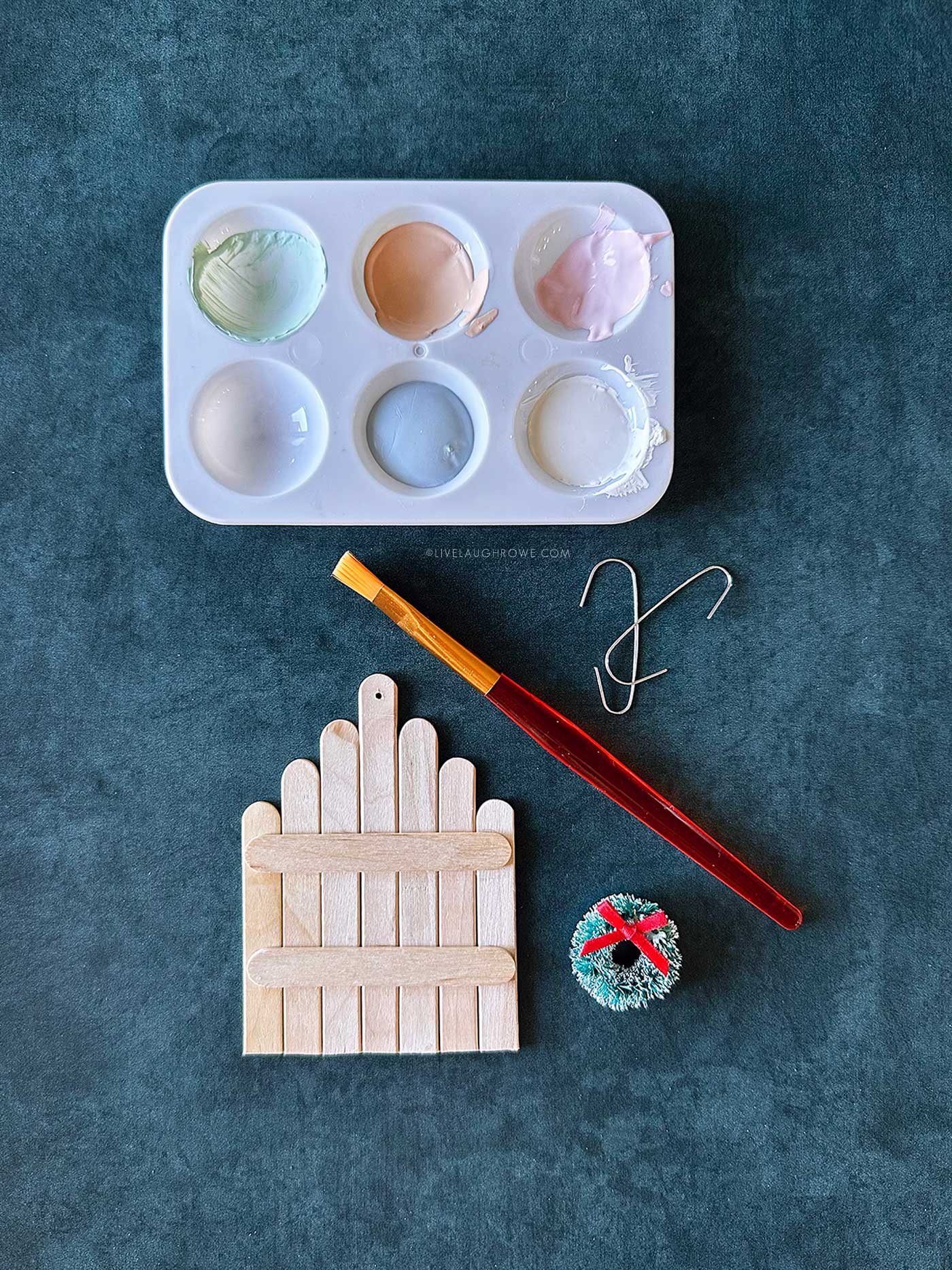 Paint palette and brush beside an unpainted popsicle stick fence ornament with a mini wreath ready for decorating.