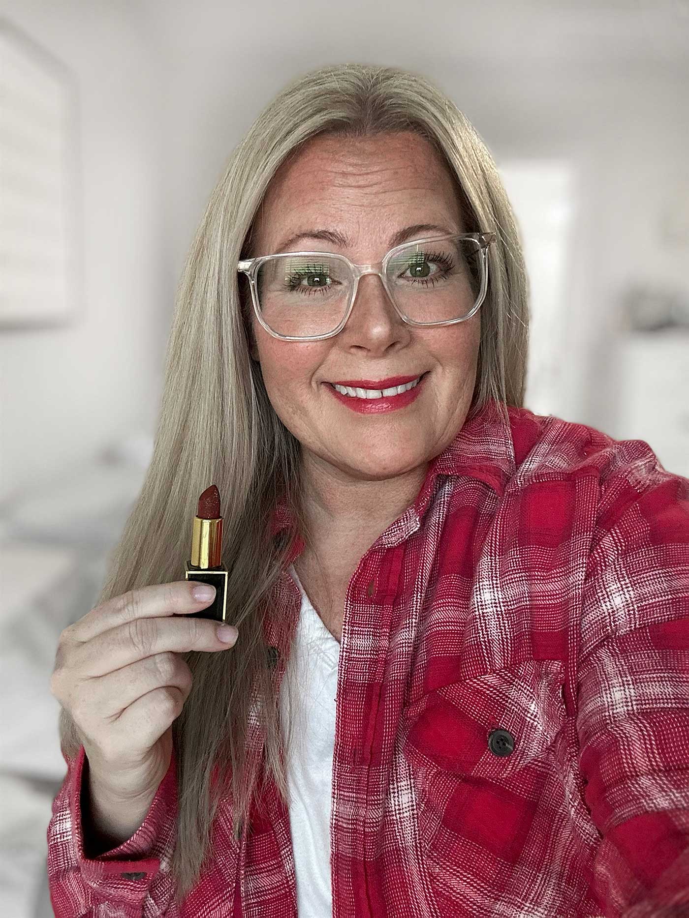 Woman wearing a red plaid shirt holding a tube of red lipstick with a soft smile in a bright room.
