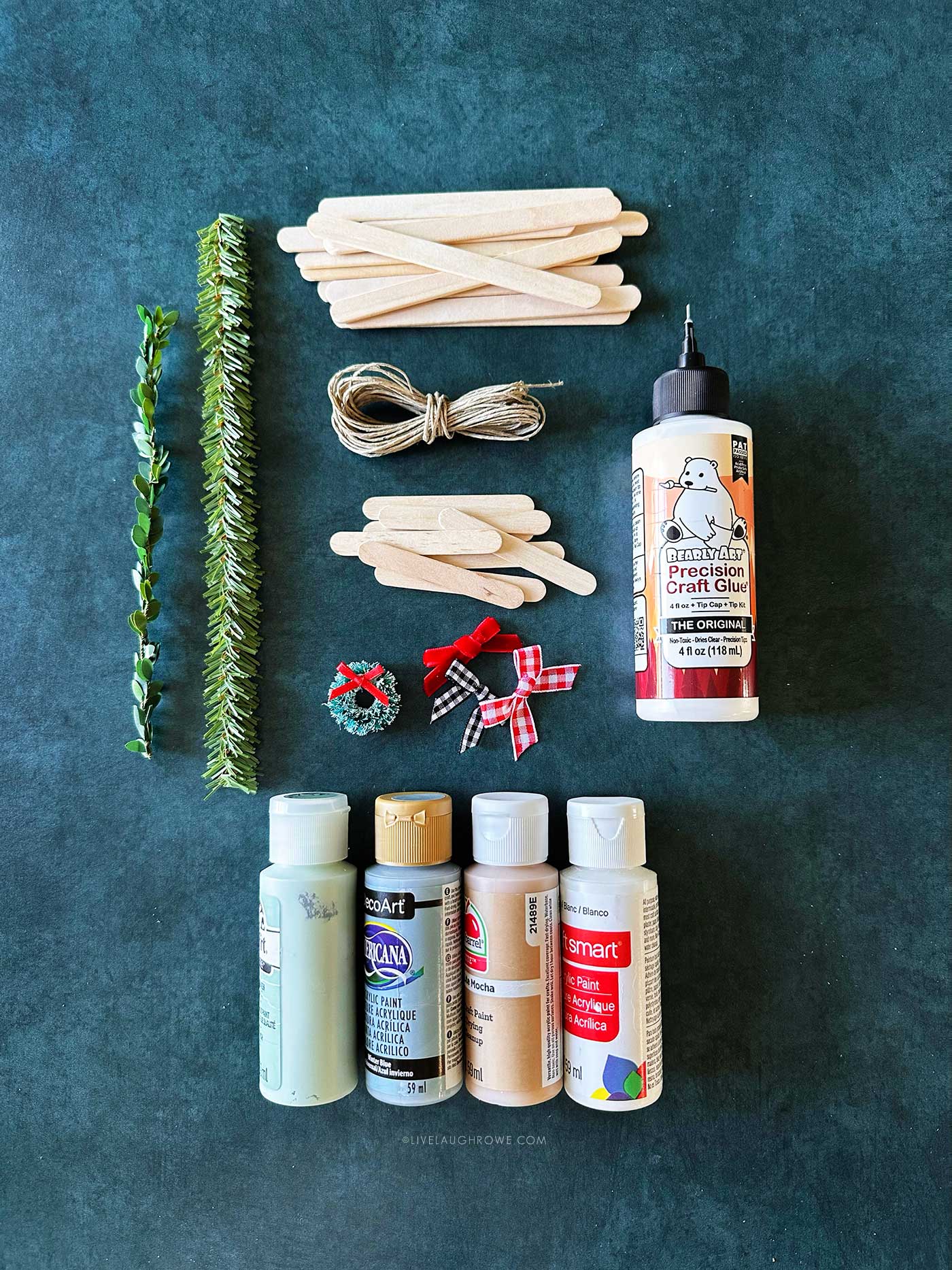 Craft supplies for making a holiday craft, including faux greenery, twine, popsicle sticks, mini wreaths, bows, paint, and craft glue.