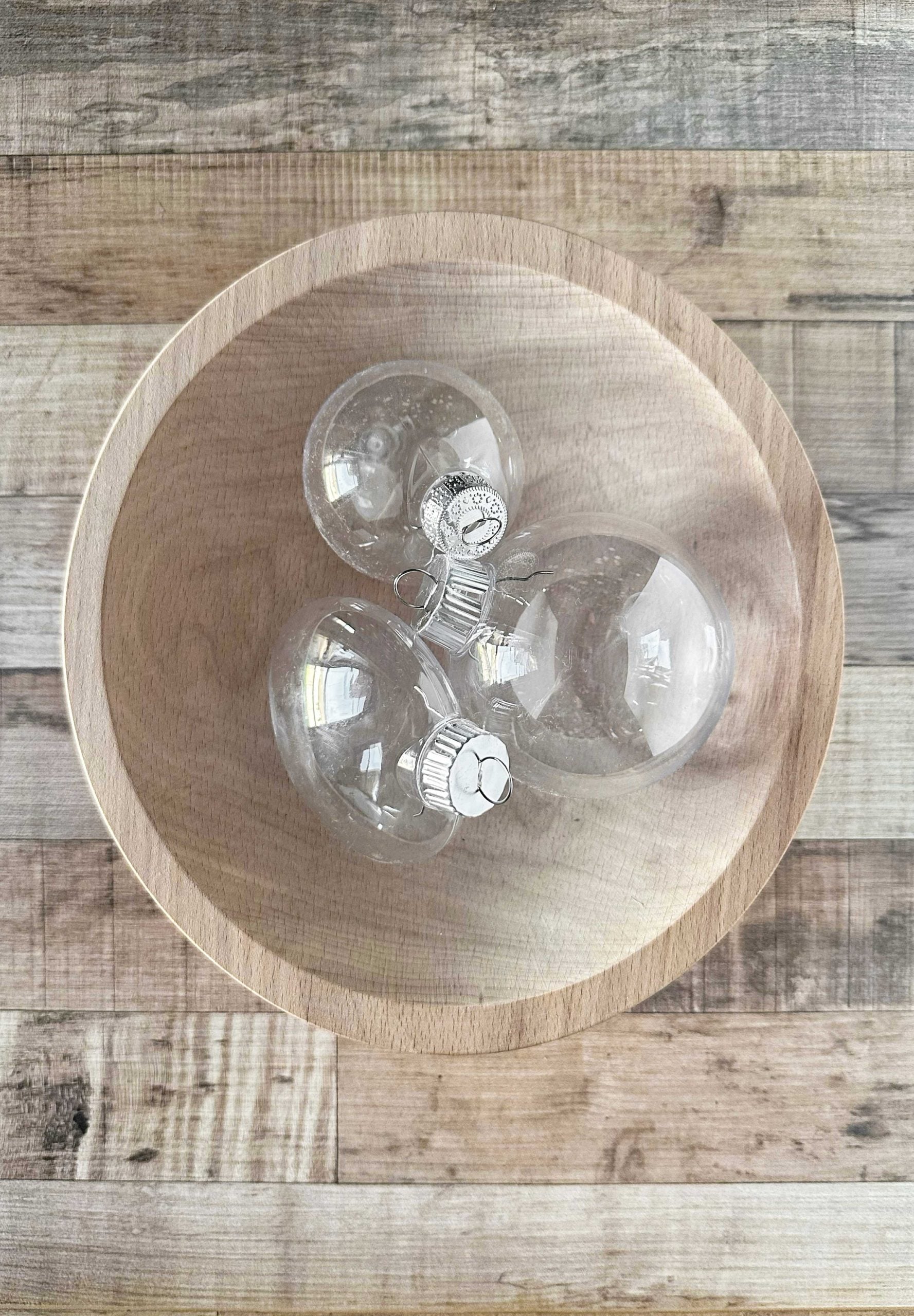 Three empty clear plastic ornaments with silver caps placed inside a wooden bowl on a woodgrain tabletop.