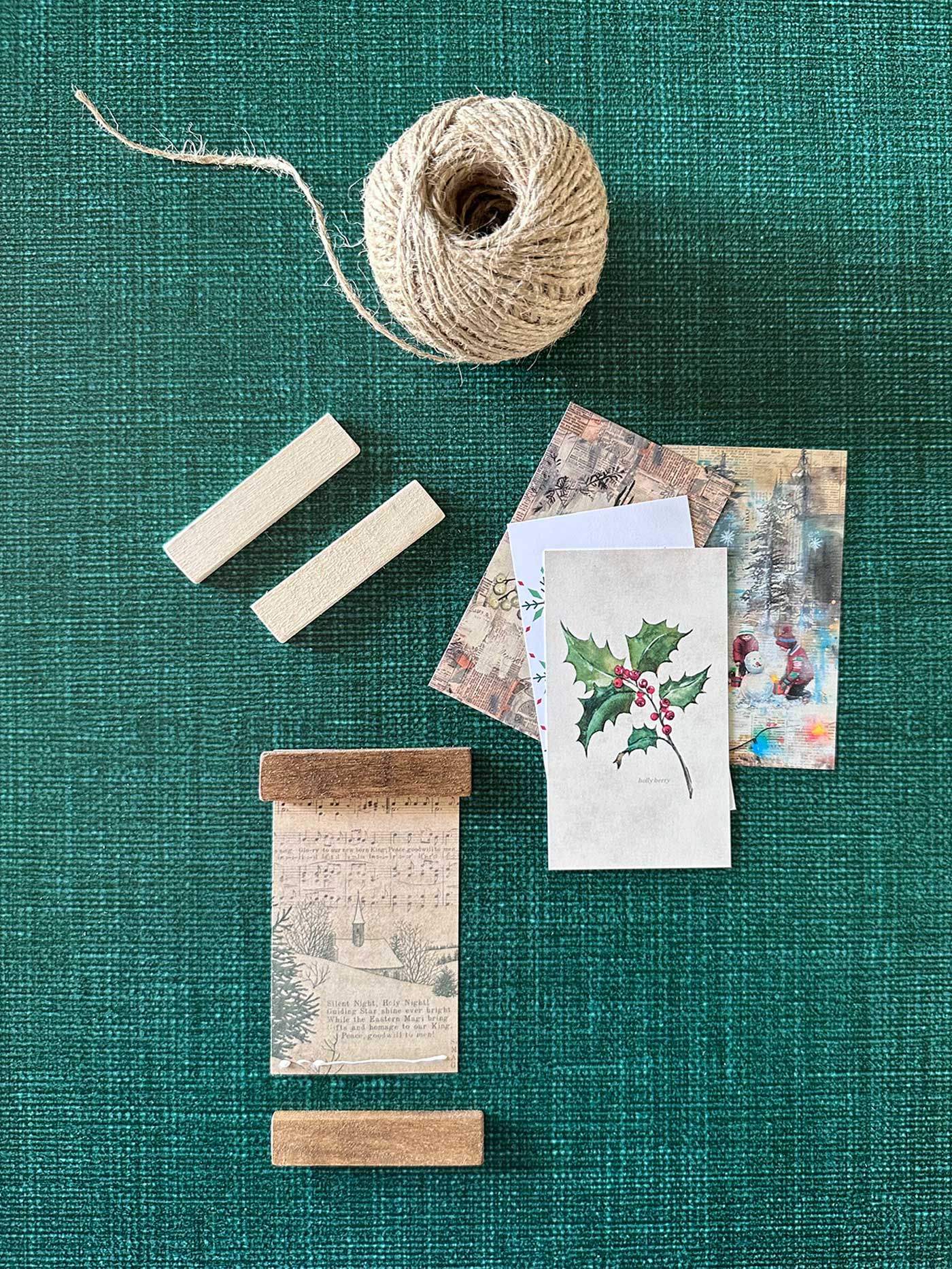Arranged materials for mini poster hanger ornaments, including twine, printed art pieces, and stained wood strips.