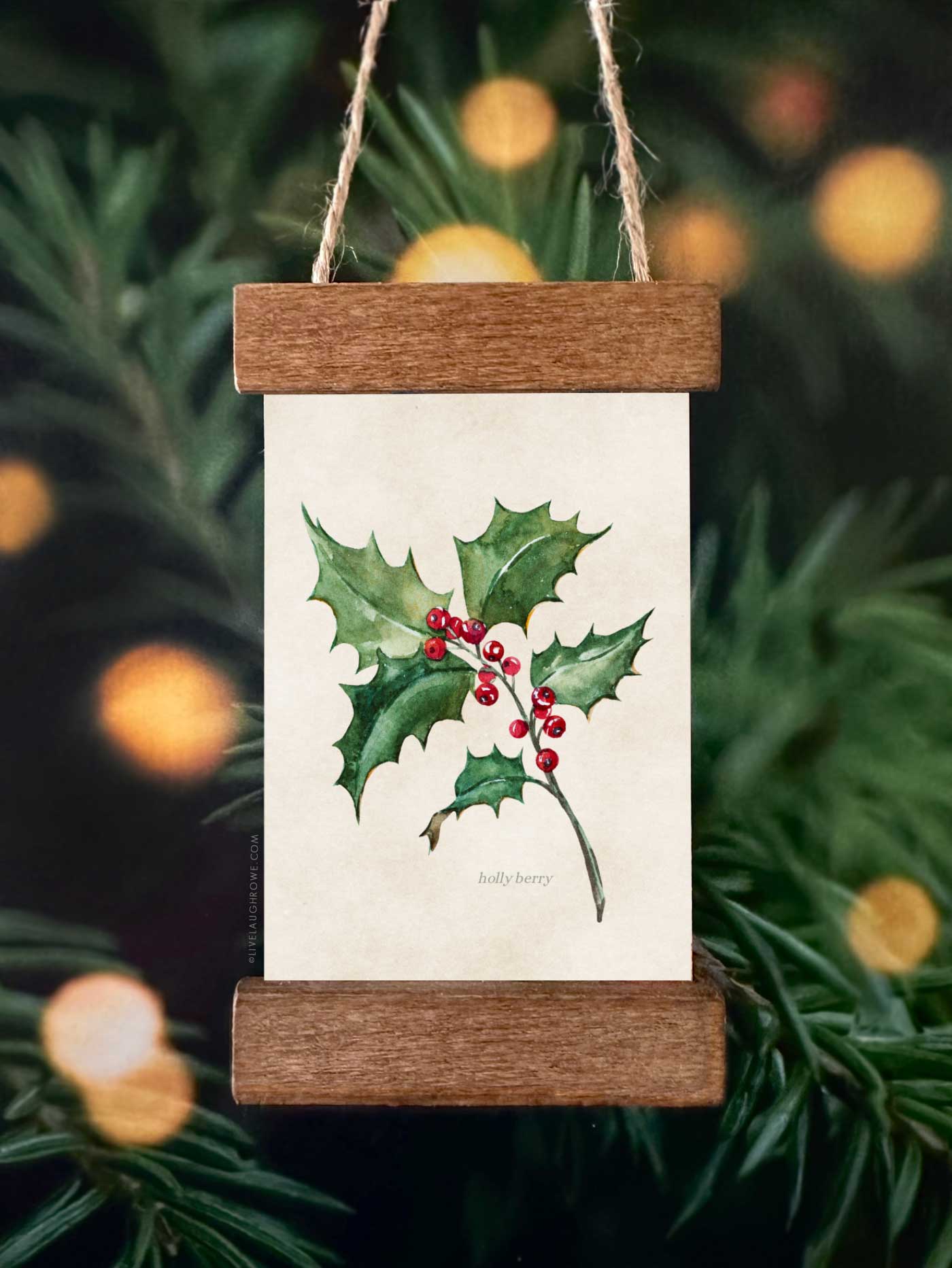 Festive ornament featuring a holly illustration displayed in a mini poster hanger ornament hanging on a Christmas tree.