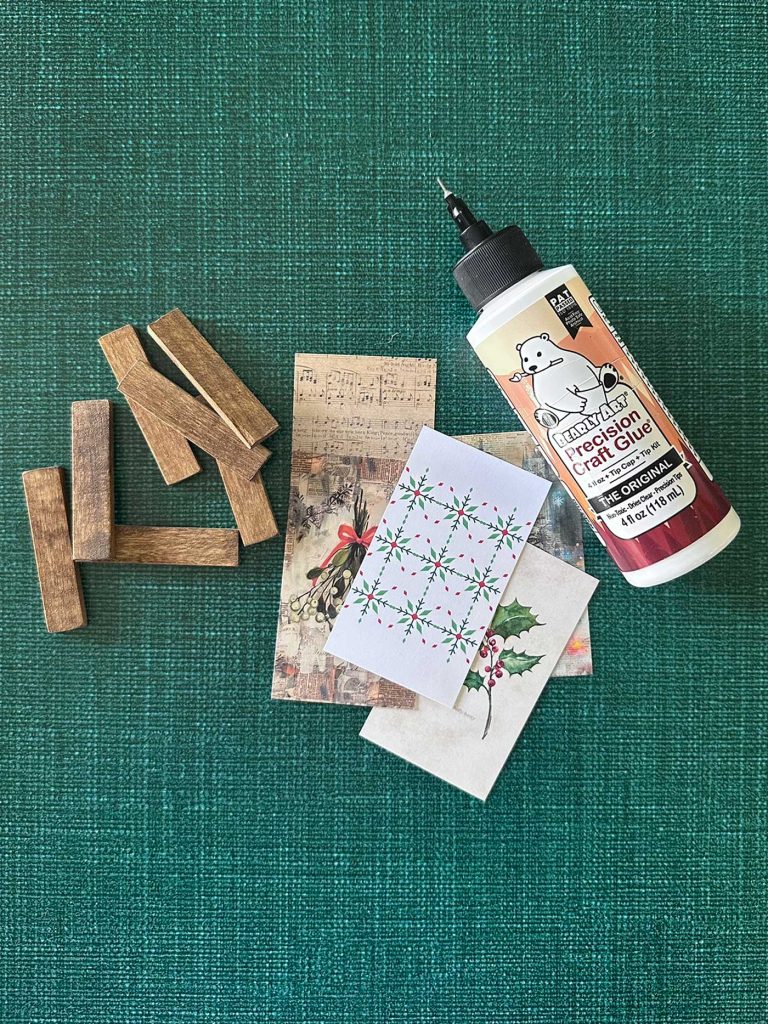 Supplies for handmade Christmas ornaments, including small wood strips, glue, and printed miniature artwork for making mini poster hanger ornaments.