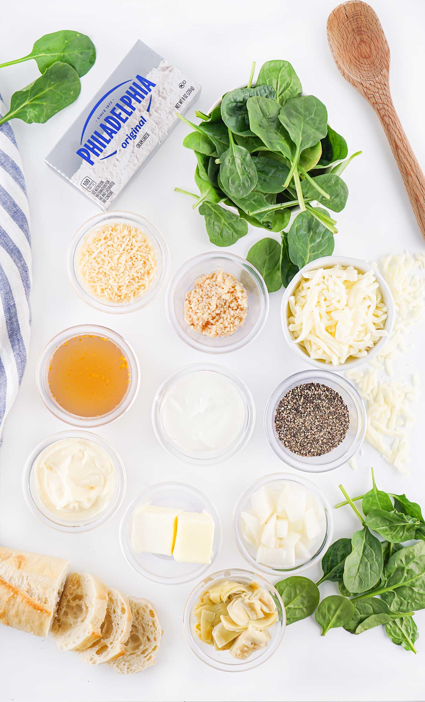 Ingredients for hot spinach dip arranged on a white surface, including spinach, cheese, artichokes, and seasonings.