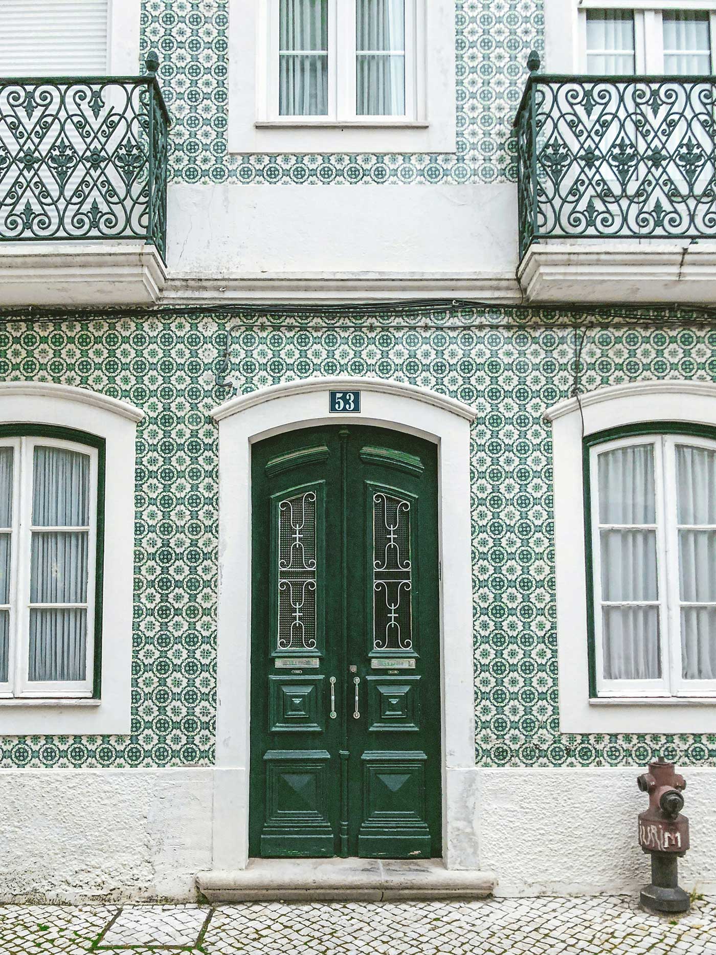 Green double doors with ornate iron details set in a white building covered in green patterned tiles, with small balconies and white-trimmed windows above.