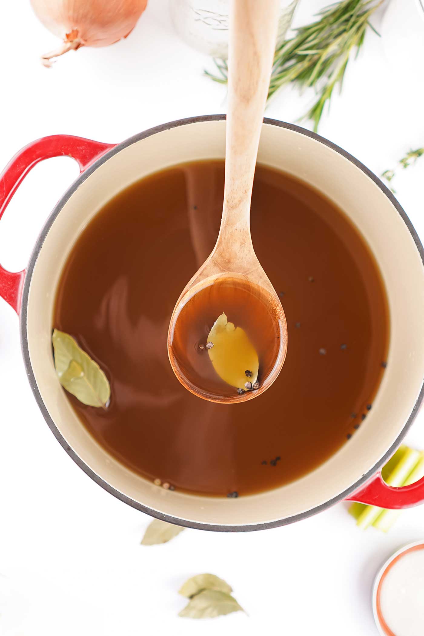 Overhead view of homemade vegetable stock simmering in a pot with bay leaves and peppercorns, wooden spoon lifting golden broth.