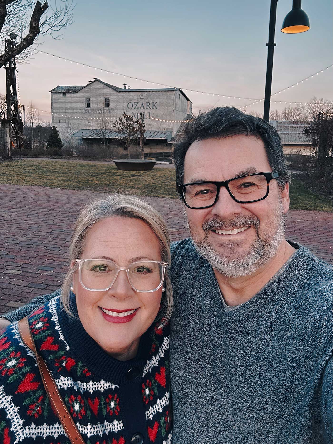 Smiling couple taking a selfie in front of The Ozark Mill at sunset in the Ozarks, with string lights and brick walkway in the background.