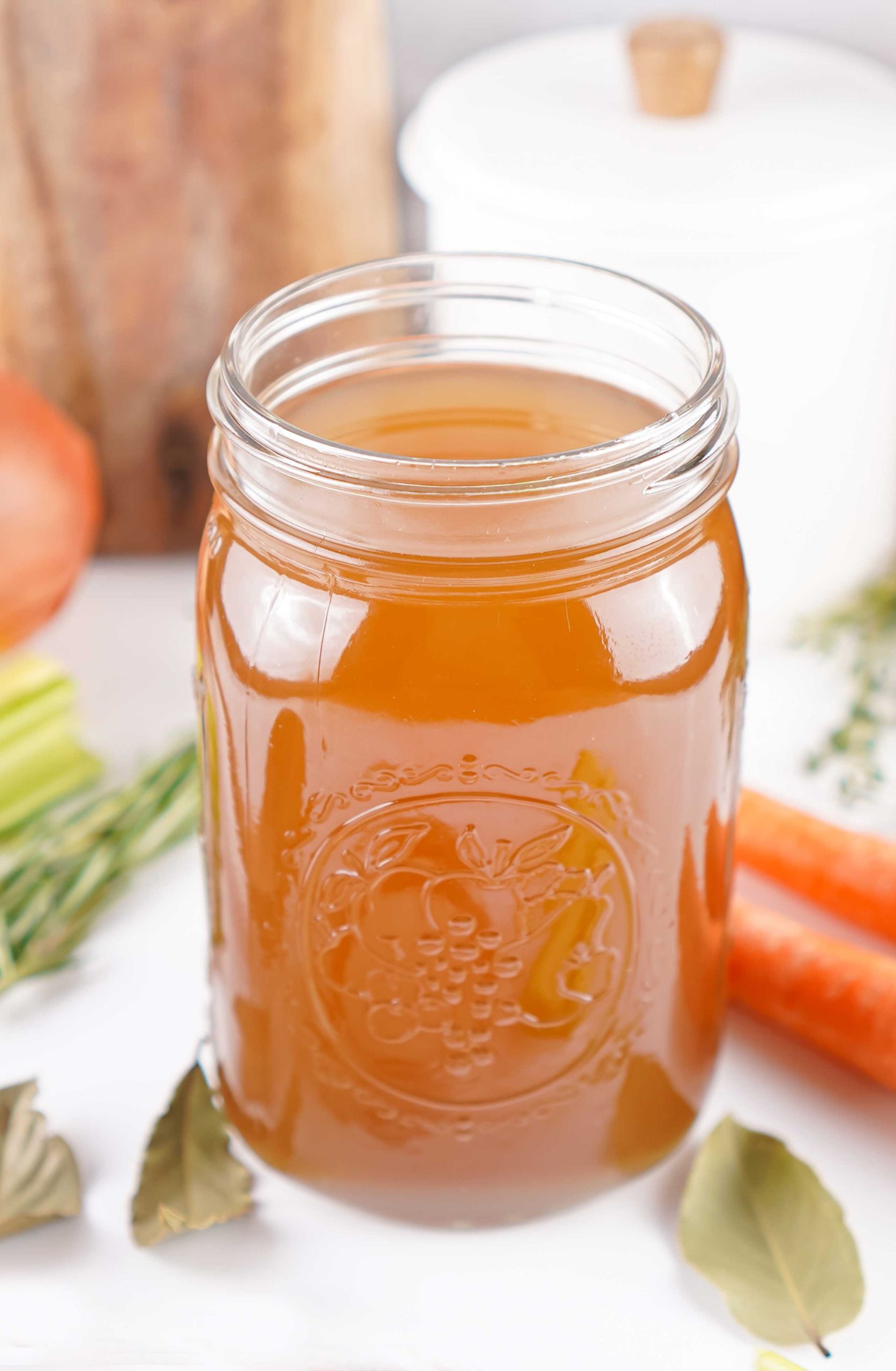 Mason jar filled with vegetable stock surrounded by fresh carrots, celery, and herbs on a white countertop.