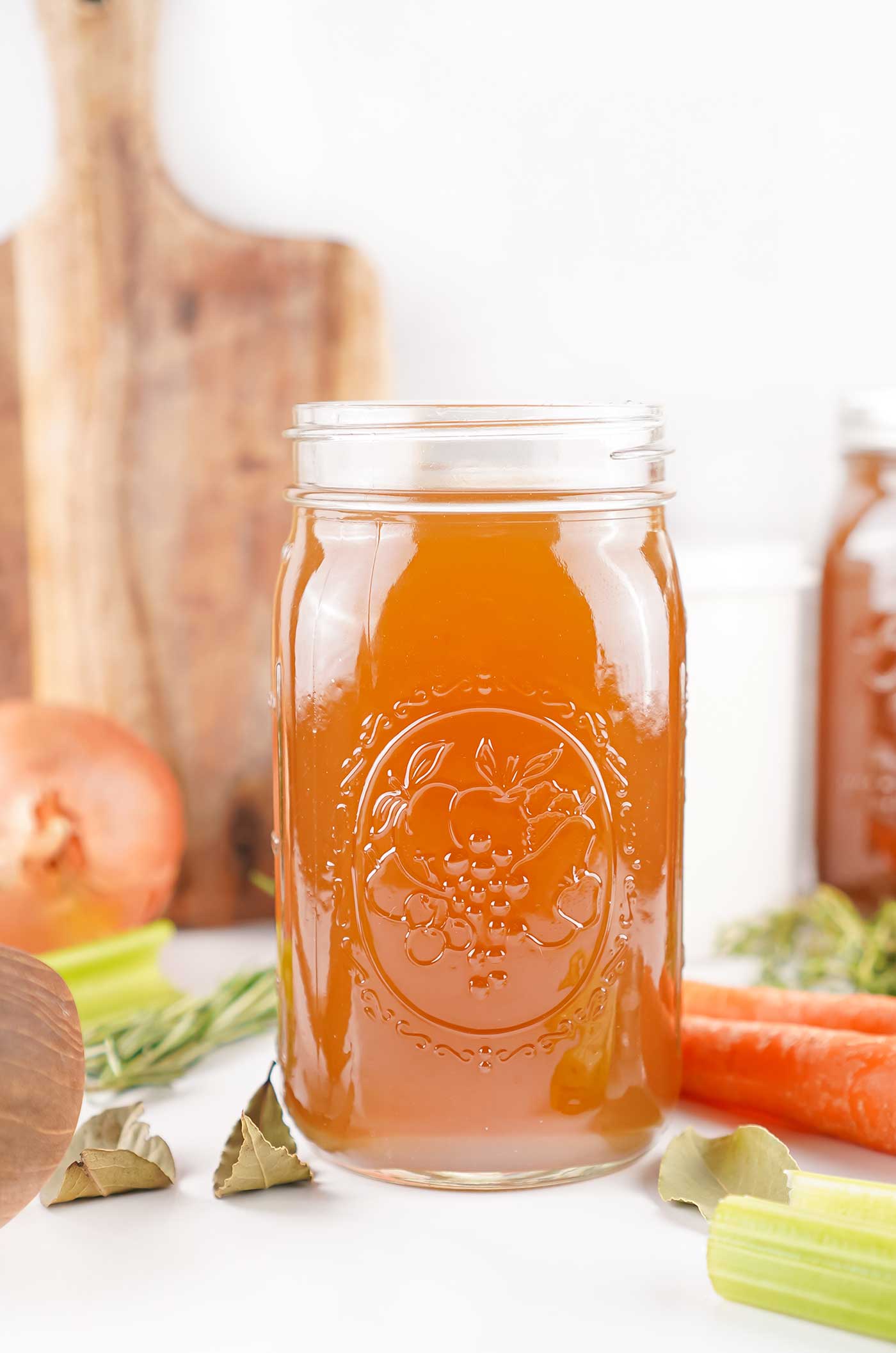 Jar of homemade stock with fresh carrots, celery, and onions on a wooden board background.