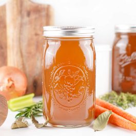 Close-up of homemade vegetable stock in a mason jar with whole carrots, celery, and bay leaves beside it.