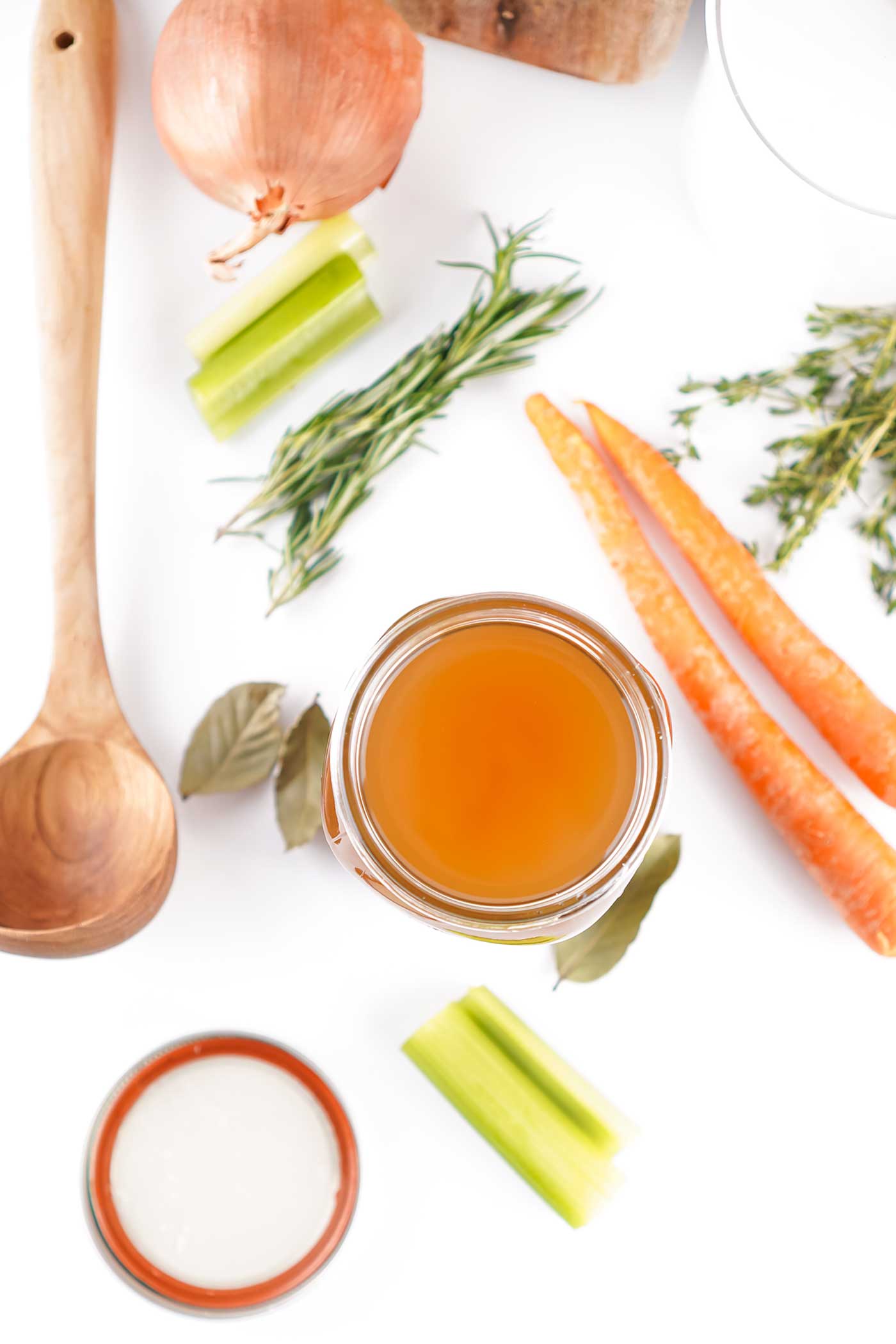 Flat lay of homemade soup base in a small bowl with wooden spoon, carrots, celery, onions, and fresh herbs