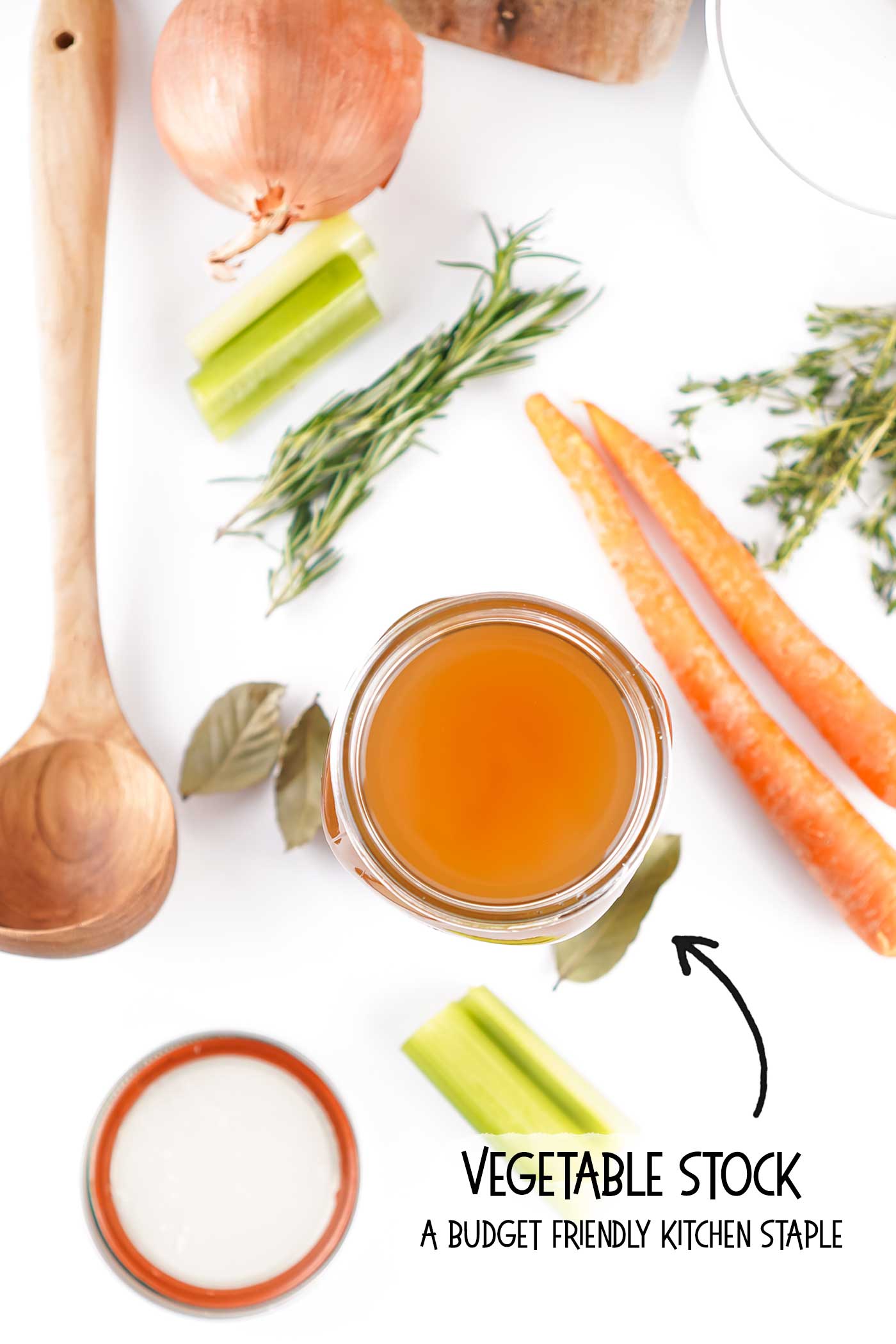 Flat lay of homemade soup base in a small bowl with wooden spoon, carrots, celery, onions, and fresh herbs