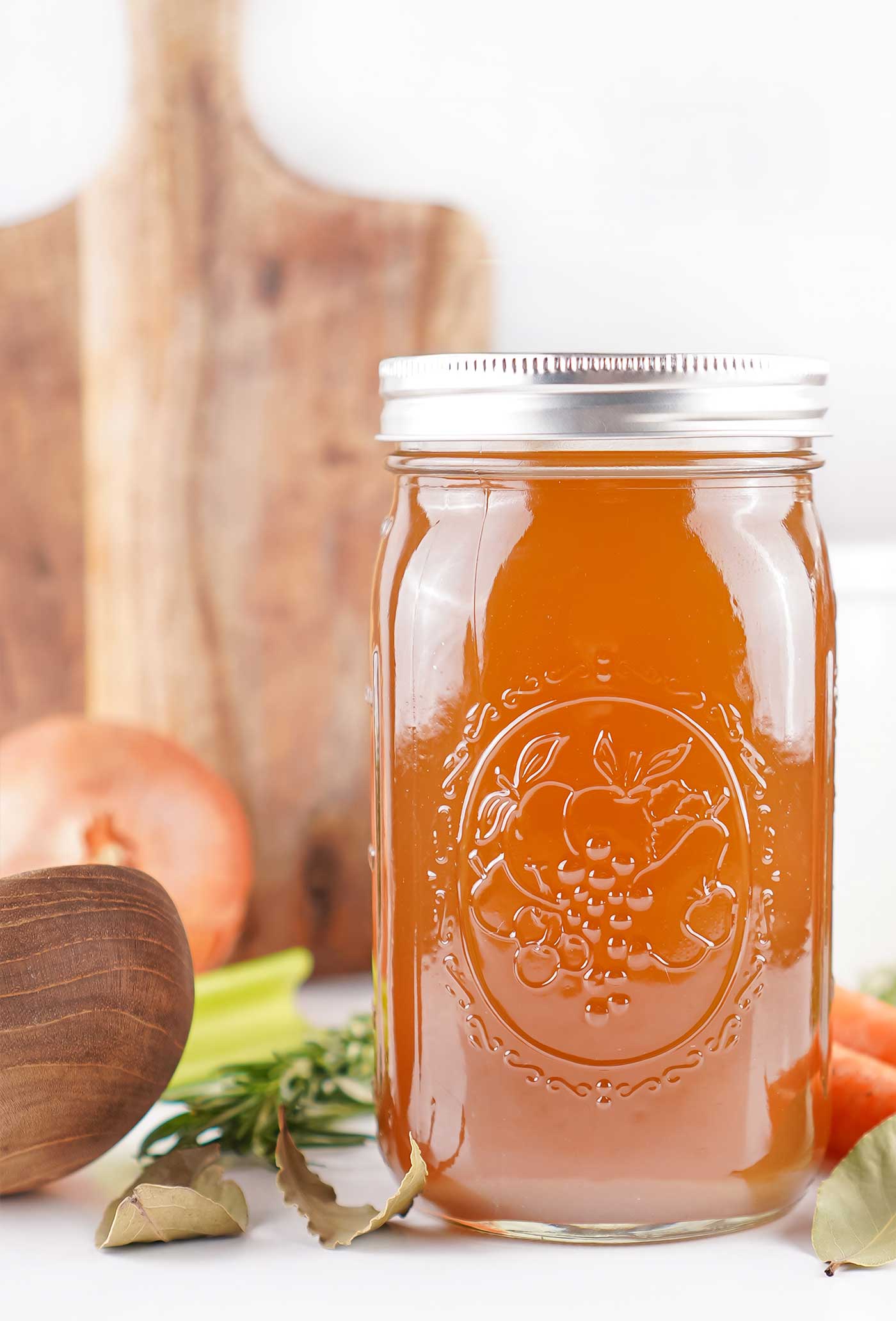 Tall mason jar of homemade soup stock with fresh vegetables and herbs styled on a rustic wooden background.