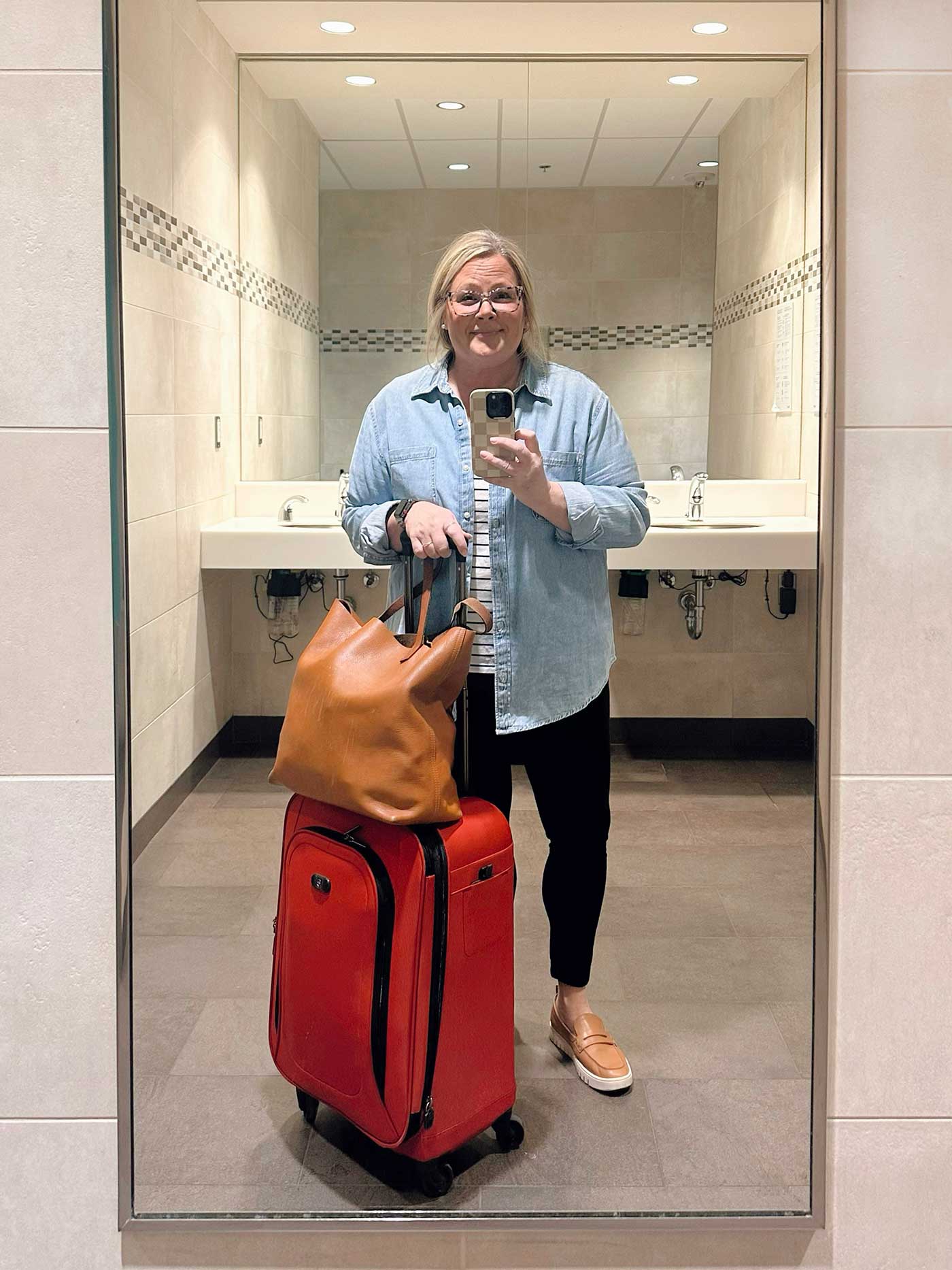 Woman taking a mirror selfie in an airport restroom with a red suitcase and tan tote bag, dressed casually for travel.