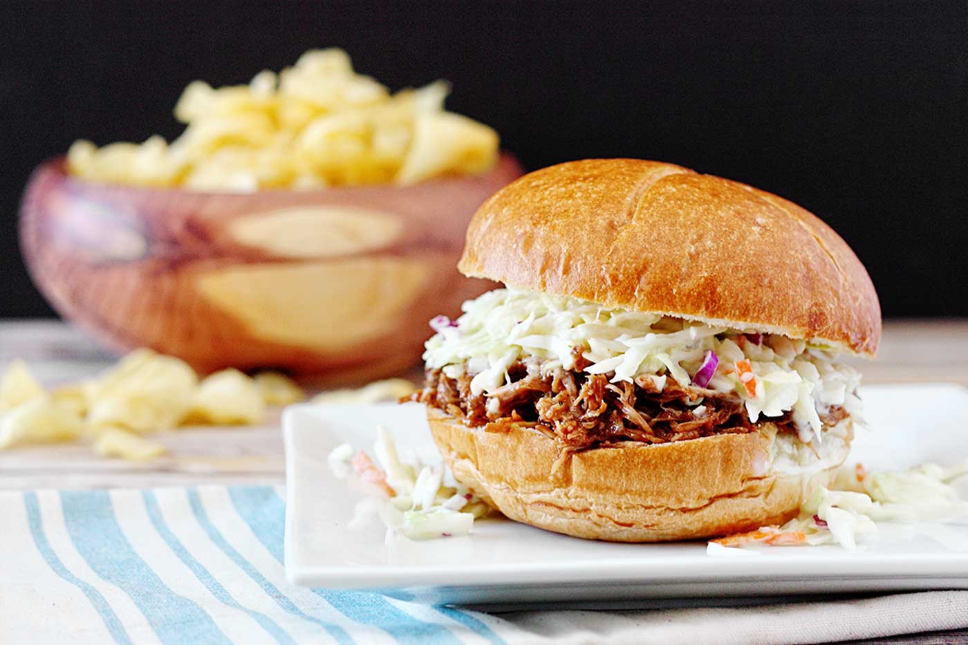 BBQ carnitas pork sandwich topped with coleslaw served on a bun with potato chips in the background