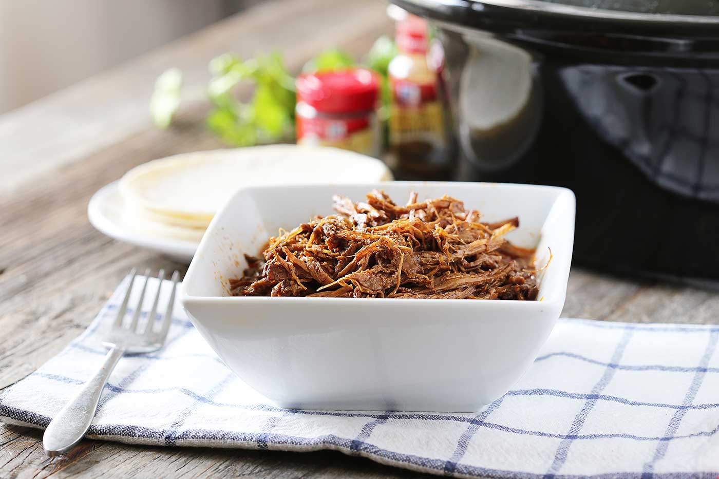 easy crockpot shredded beef served in a white bowl with tortillas and seasoning in the background