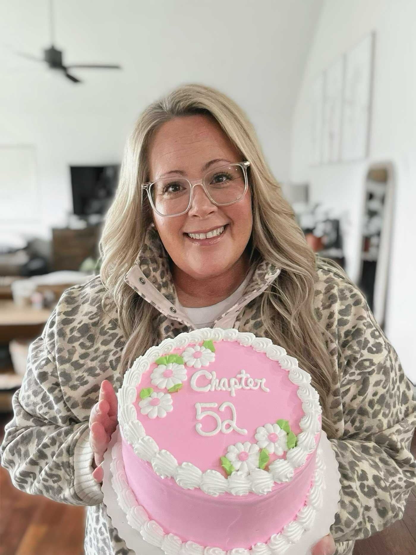 Woman smiling and holding a pink birthday cake decorated with flowers and the words Chapter 52.