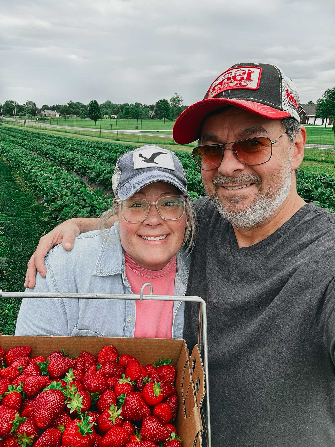Woman and man smiling while holding a basket of freshly picked strawberries at a strawberry farm on a cloudy day.